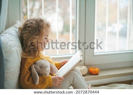 Little cute curly haired girl in knitted pullover sits in front of the window. Cozy home. Warm autumn.