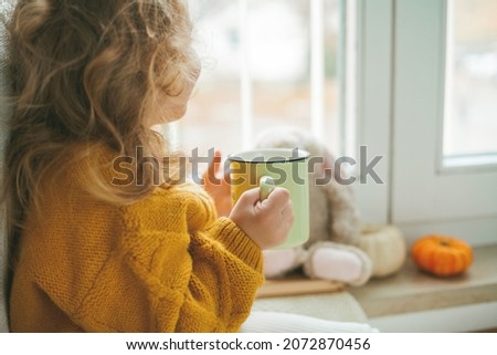 Little cute curly haired girl in knitted pullover is drinking tea and eating cookies in front of the window. Cozy home. Warm autumn. Mood.