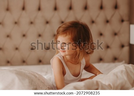 Cute toddler girl playing in bed with white linen. Cozy beroom interior in neutral beige colors. Good morning concept 