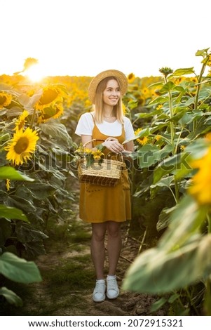 A smiling woman is carrying a basket of flowers. She is standing on a field of sunflowers and smiling. Sunset.