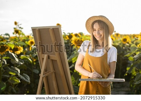 A woman is standing in a field of sunflowers and drawing a picture. She's wearing a straw hat. The canvas is standing on an easel. She is smiling and looking at the camera.
