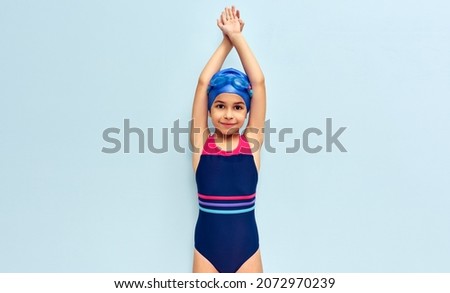 Horizontal image of a cute little girl in goggles, a swimsuit, and a blue swimming cap get ready before diving isolated blue studio background. Kid in swimsuit making swimming gesture before the dive.