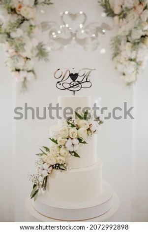 White wedding cake in three tiers decorated with white peonies and cotton on a wall background with a picture of swans