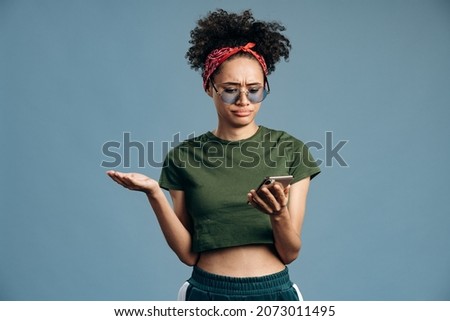 Multiracial woman holding smartphone and feeling disappointed of something. Indoor studio shot isolated on blue background 