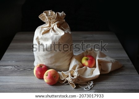 There are two bags on the wooden table. One bag is full, the other lies empty next to it. Next to the bags are three ripe large red apples. An image with selective focusing.