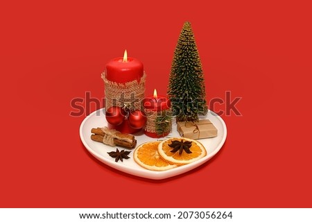 White plate with a red candle and dried oranges and a pine cone. Mini tree. On a red background.