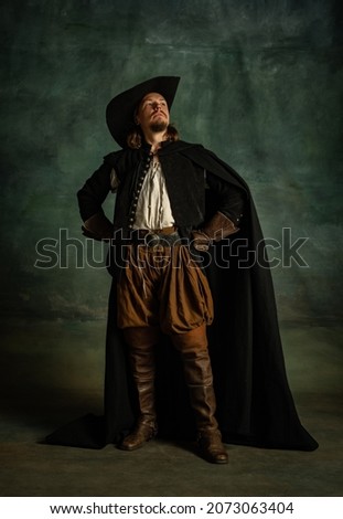 Portrait of genteel brutal man, medeival pirate in hat and cloak with sword isolated over dark background. Combination of medeival and modern styles. Concept of history. Copyspace for ad.