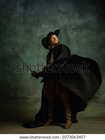 Portrait of fearless man, medeival pirate in hat and cloak with sword isolated over dark background. Combination of medeival and modern styles. Concept of history. Copyspace for ad.