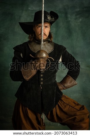 Portrait of brutal man, medeival pirate in vintage costume holding sword isolated over dark background. Ready to fight. Combination of medeival and modern styles. Concept of history. Copyspace for ad.
