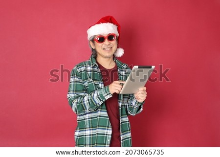 The senior Asian man wears green plaid shirt with Santa Claus hat standing on the red background.