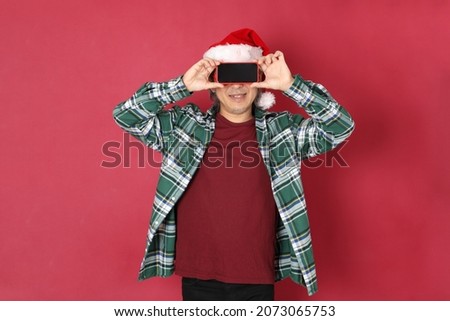 The senior Asian man wears green plaid shirt with Santa Claus hat standing on the red background.