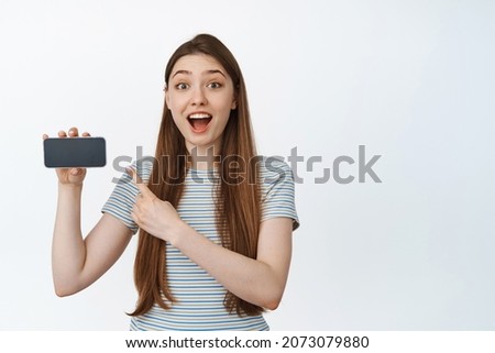 Surprised and happy woman pointing finger at horizontal smartphone screen, smiling amazed, showing app advertisement, website on mobile phone, white background