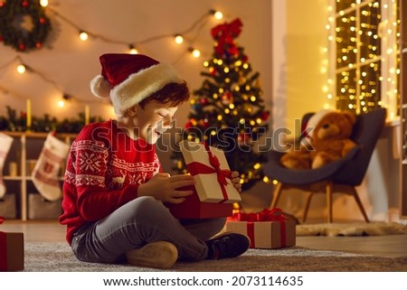 Just believe in magic. Happy surprised kid in Santa hat unpacks New Year gift. Luminous glow lights up little boy's face as he opens his wonderful present sitting in the living-room on Christmas night