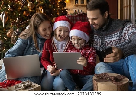 Addicted to technology overjoyed family couple with kids watching funny Christmas cartoons or comedian movies online, holding electronic gadgets in hands, New Year winter holiday celebration. Royalty-Free Stock Photo #2073130589
