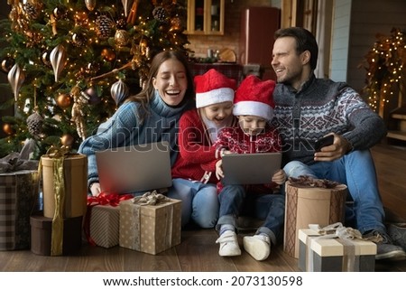 Technology addicted overjoyed young family couple watching funny cartoons online or playing games on touchpad with happy sincere small adorable children siblings, unwrapping Christmas gifts at home.
