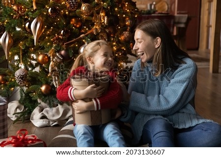 Overjoyed little kid girl cuddling wrapped in box present, feeling excited of getting wished Christmas gift, sitting with affectionate happy mother near decorated festive tree in living room.