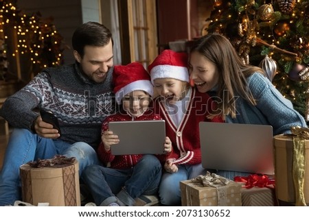 Happy young parents sitting on floor near decorated Christmas tree, watching funny comedian movie or cartoons with adorable small children on digital tablet, unpacking gifts together in living room.