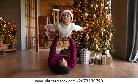 Wide banner panoramic view of happy little biracial boy have fun play with loving young father at home. African American dad lying on floor near Christmas tree engaged in funny game with small son.