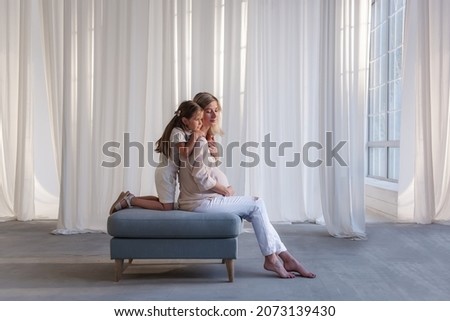 Little daughter hugs pregnant mother in white studio. Girl with young woman sit on chair against the background of floating airy fabrics. Easy, healthy pregnancy, the joy of motherhood. Copy space