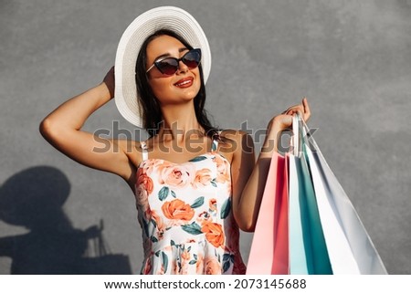 Stylish beautiful happy brunette in hat, holding shopping bags for Black Friday, standing outdoors on isolated gray background, Copy space, Shopping concept, Black Friday, clearance sale, discounts