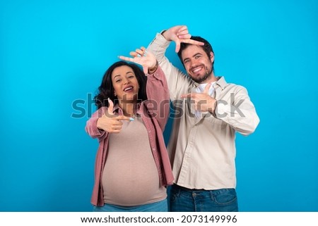 young couple expecting a baby standing against blue background making finger frame with hands. Creativity and photography concept.