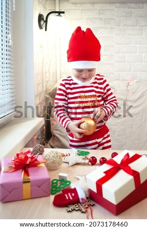 A small child in a Santa Claus hat is holding a large golden Christmas tree toy. Christmas presents are on the table.