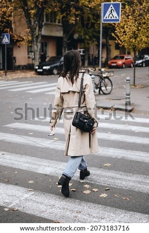 Girl cross the street at a pedestrian crossing. Trendy street style photography. The girl dressed in a beige trench coat walks through the autumn city