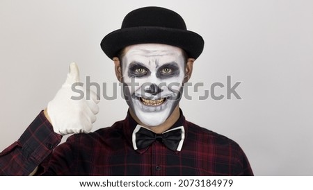 A horrible man in clown makeup smiles and shows a thumb up. A scary clown looks at the camera and laughs terribly.