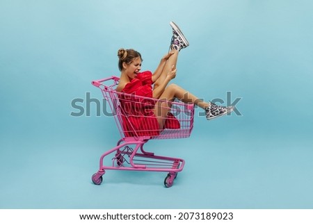 Full growth energetic young caucasian girl sits in shopping trolley pictured in blue studio. Crazy slender model with blond tied hair lifts her leg up, wearing red dress.