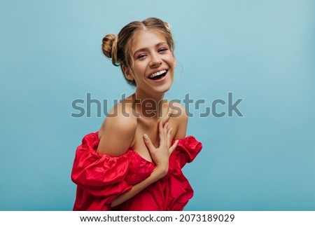 Close-up fair-skinned young lady laughing hard while holding chest with one hand on blue background. Pretty model in red summer outfit with gathered blond hair. Emergence of positive emotion, concept Royalty-Free Stock Photo #2073189029