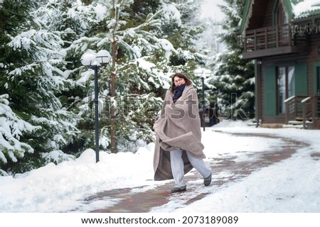 A beautiful brunette middle-aged happy woman in glasses covered with a warm beige knit blanket walking the paved path near the house with the snowy forest on her back 
