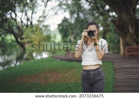 Female photographer standing and using millorless camera taking photo looking at camera with fresh green park background.