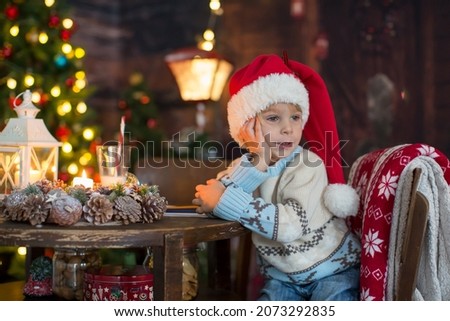 Cute toddler child, boy in a Christmas outfit, playing in a wooden cabin on Christmas, decoration around him. Child reading book and drinking tea