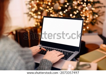 woman work on laptop computer with blank display screen with mockup copy space. home interior decorated for Christmas celebration with Christmas tree and garland lights. Royalty-Free Stock Photo #2073357497