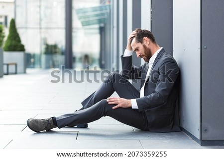 Sad depressed entrepreneur in formal suit worker man sitting near outdoors street near modern office business center. Upset male businessman lost job due financial crisis employee has problem. outside Royalty-Free Stock Photo #2073359255
