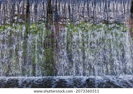Fragment of an artificial waterfall formed by the concrete dam, covered with green moss in park

