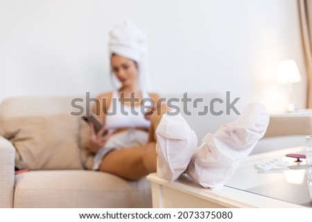 Close-up of woman's feet wrapped in moisturizing socks during home pedicure. Young happy woman texting on mobile phone while using moisturizing socks on her feet after having pedicure at home.