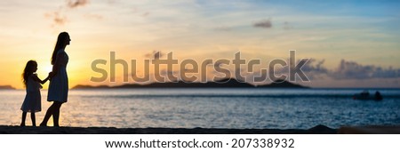 Silhouettes of mother and daughter walking along tropical beach during sunset, panorama with copy space perfect for banners
