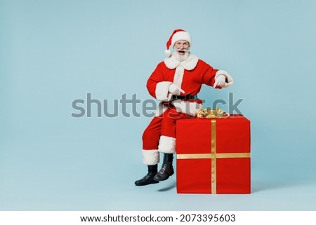 Full body old Santa Claus man in Christmas hat red suit clothes sitting point on big gift present box isolated on plain blue background studio. Happy New Year 2022 celebration merry ho x-mas concept