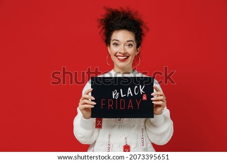 Young smiling caucasian happy female costumer woman 20s wear white knitted sweater with tags sale hold in hand card sign with balck friday title text isolated on plain red background studio portrait