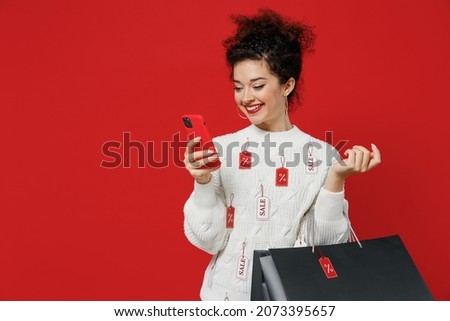 Young smiling happy female costumer woman wear white knitted sweater with tags sale hold package bags with purchases after shopping using mobile cell phone isolated on plain red background studio.
