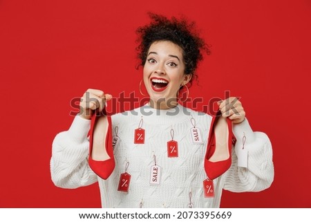 Young surprised happy caucasian female costumer woman 20s wear white knitted sweater with tags sale in store showroom hold high heels shoes look camera isolated on plain red background studio portrait