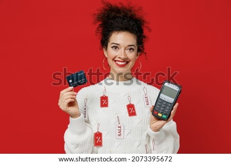 Young happy female costumer woman 20s in white knitted sweater with tags sale hold wireless modern bank payment terminal to process acquire credit card paymentsisolated on plain red background studio