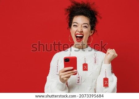 Young happy female costumer woman 20s wear white knitted sweater with tags sale in store showroom hold in hand use mobile cell phone do winner gesture isolated on plain red background studio portrait