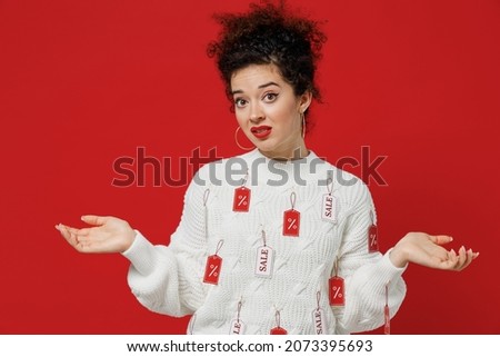 Young doubtful costumer woman 20s wear white knitted sweater with tags sale in store showroom spread hands shrugging shoulders looking puzzled, have no idea isolated on plain red background studio.