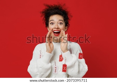 Young promoter female costumer woman 20s wear white knitted sweater with tags sale in store showroom scream hot news about sales discount with hands near mouth isolated on plain red background studio