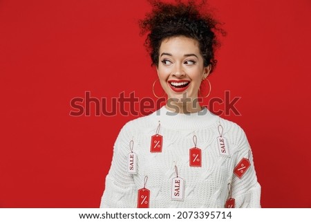 Young smiling cheerful satisfied happy female costumer woman 20s wear white knitted sweater with tags sale in store showroom looking aside on workspace isolated on plain red background studio portrait