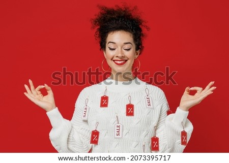 Young female costumer woman in white knitted sweater with tags sale in store showroom hold spreading hands in yoga om aum gesture relax meditate try calm down isolated on plain red background studio