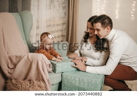 Young family husband, wife and daughter sitting on chair at home near the Christmas tree. Dad and mom hold the baby, hugs, joy, happiness, opening gifts. Decorated house, quarantine coronavirus.