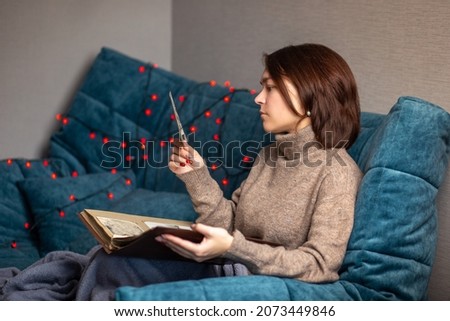 a brunette girl sits on a blue sofa with an old photo album, memories, important photos, the transience of life, deceased loved ones, longing for loved ones, longing for isolation, depression, joy of  Royalty-Free Stock Photo #2073449846